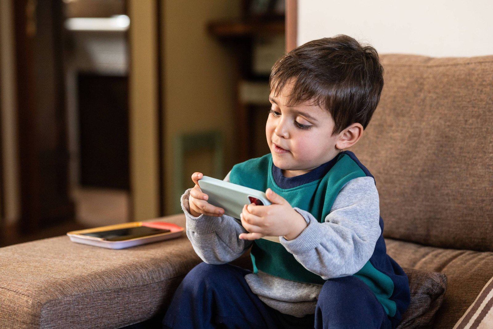 Um menino pequeno está sentado em um sofá marrom, segurando um smartphone verde. Ele está vestido com uma camiseta azul e um suéter cinza. Ao lado dele, há um caderno e outro dispositivo móvel. O ambiente parece ser uma sala de estar com iluminação suave.