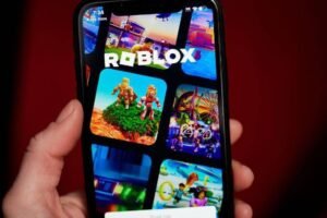 A imagem mostra um smartphone sendo segurado por uma mão, exibindo a tela do aplicativo Roblox. A tela apresenta várias miniaturas de jogos, com cores vibrantes e personagens em diferentes cenários. O logotipo do Roblox está em destaque na parte superior da tela.