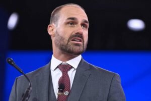 Um homem com cabelo curto e barba está falando em um microfone. Ele usa um terno cinza escuro com uma gravata vermelha e uma camisa branca. O fundo é desfocado, mas parece ser um ambiente de conferência ou palestra.