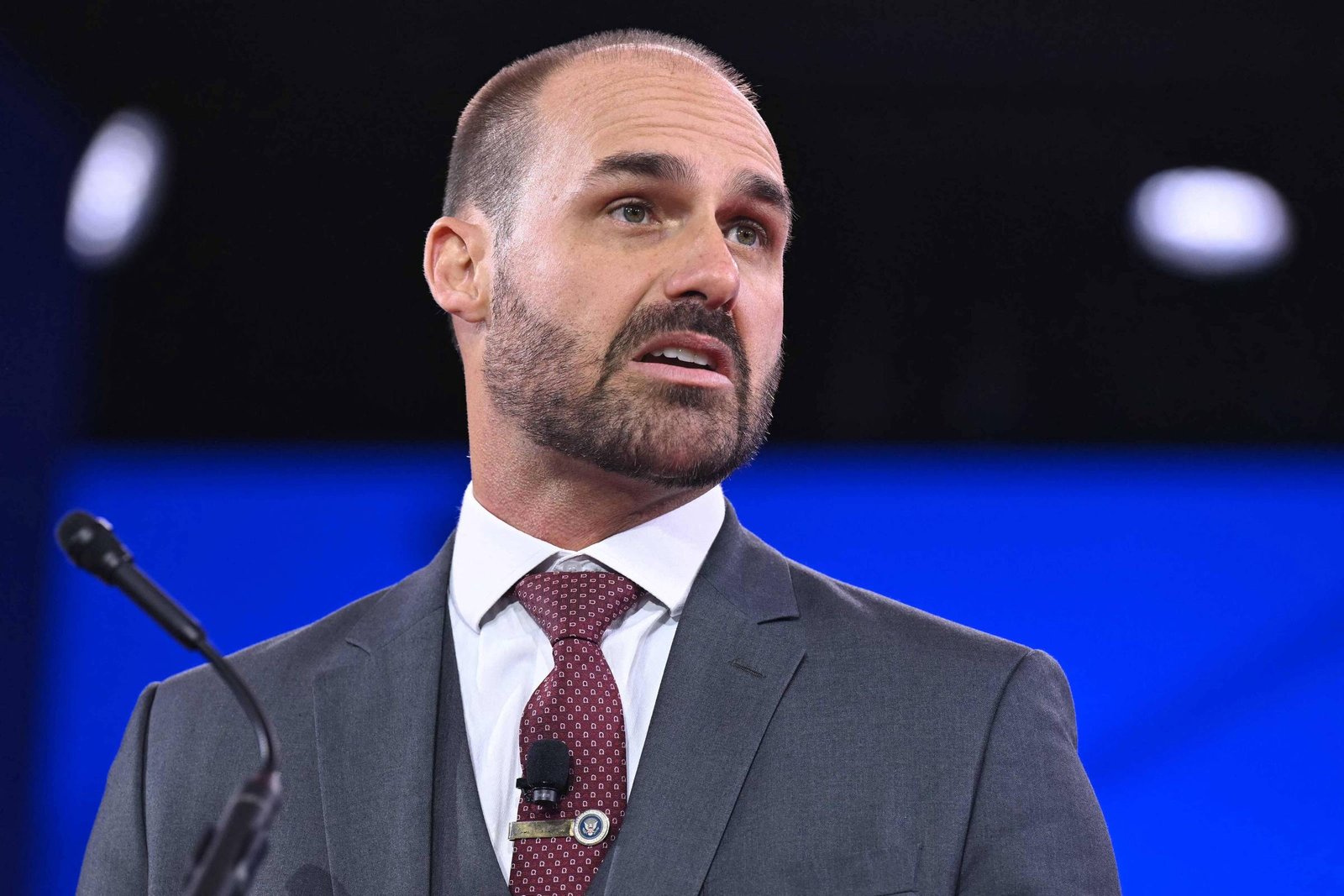 Um homem com cabelo curto e barba está falando em um microfone. Ele usa um terno cinza escuro com uma gravata vermelha e uma camisa branca. O fundo é desfocado, mas parece ser um ambiente de conferência ou palestra.