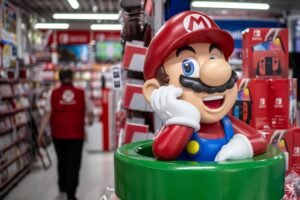 A imagem mostra uma estátua do personagem Mario, da Nintendo, saindo de um tubo verde. Ele está sorrindo e apoiando a cabeça com a mão. Ao fundo, há prateleiras com produtos de jogos e um funcionário vestido com uma camisa vermelha, caminhando pela loja.