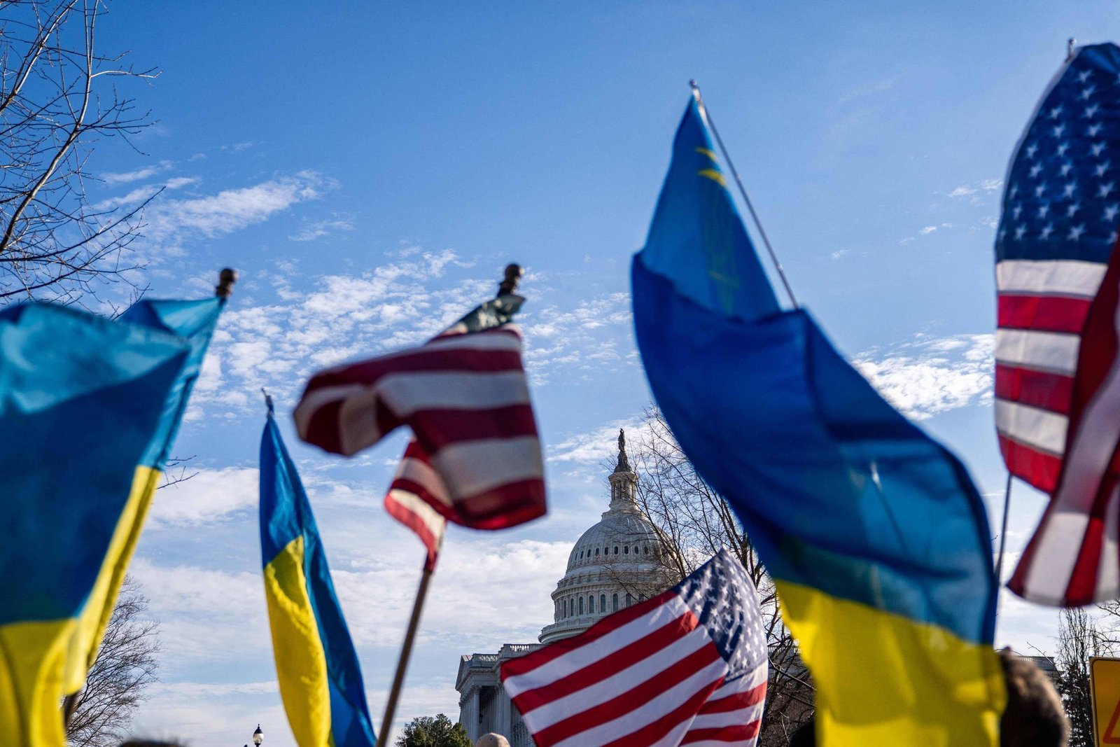 A imagem mostra um grupo de pessoas segurando bandeiras da Ucrânia e dos Estados Unidos em frente ao Capitólio. O céu está claro com algumas nuvens. O edifício do Capitólio é visível ao fundo, parcialmente coberto pelas bandeiras.