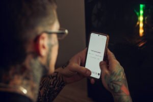 A imagem mostra uma pessoa com tatuagens visíveis nos braços, usando óculos, segurando um smartphone com a tela em branco. Ao fundo, há um objeto com luzes coloridas, possivelmente um computador ou um dispositivo eletrônico.