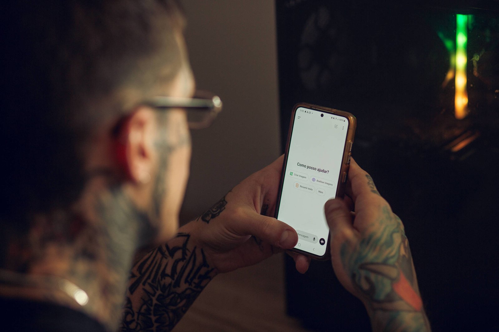 A imagem mostra uma pessoa com tatuagens visíveis nos braços, usando óculos, segurando um smartphone com a tela em branco. Ao fundo, há um objeto com luzes coloridas, possivelmente um computador ou um dispositivo eletrônico.