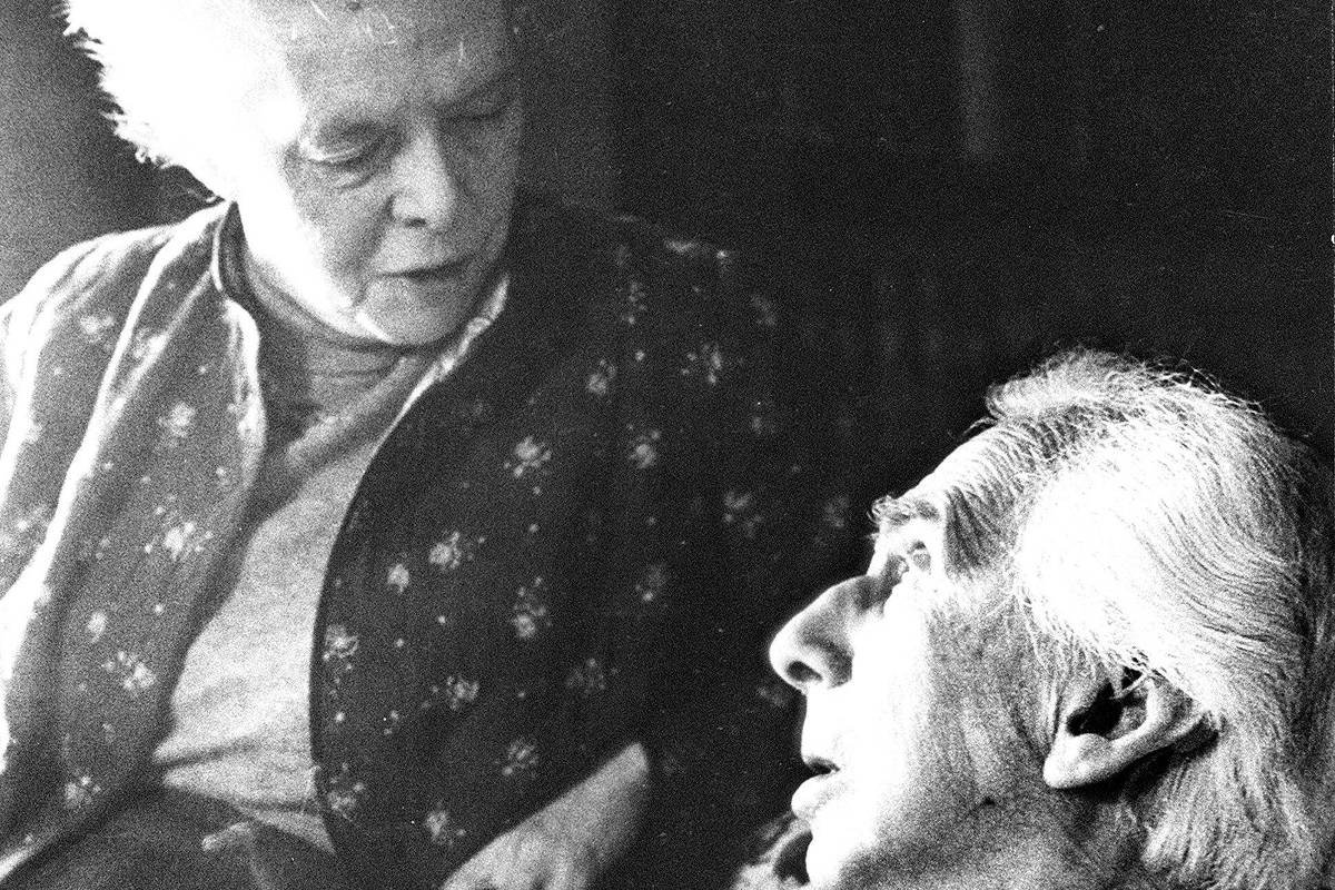 A imagem em preto e branco mostra duas pessoas idosas em uma conversa. A mulher, com cabelo claro e preso, está olhando para o homem, que tem cabelo grisalho e está sentado. Ele parece estar falando, enquanto ela escuta atentamente. Ambos estão vestidos de forma formal, com a mulher usando um suéter e o homem um colete e paletó. O fundo é desfocado, sugerindo um ambiente interno.