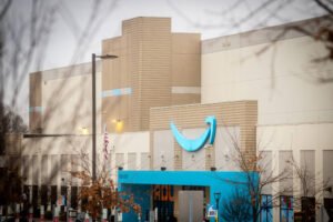 A imagem mostra a fachada de um grande centro de distribuição. O edifício é de cor clara, com detalhes em azul. Na parte frontal, há um grande símbolo em forma de sorriso, que é característico de uma empresa de comércio eletrônico. Árvores sem folhas estão visíveis na frente, sugerindo que a foto foi tirada em uma estação mais fria. O céu está nublado.