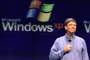 A imagem mostra um homem sorridente em um palco, usando um suéter azul e segurando um microfone. Ao fundo, há um grande logotipo do Windows XP, com as palavras