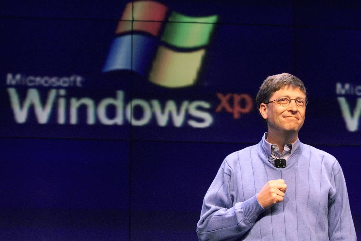 A imagem mostra um homem sorridente em um palco, usando um suéter azul e segurando um microfone. Ao fundo, há um grande logotipo do Windows XP, com as palavras