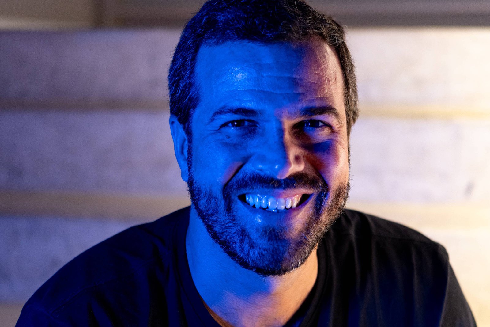 Um homem com barba e cabelo curto, vestindo uma camiseta preta, está sentado em uma escada. Ele sorri amplamente, e a iluminação azul destaca seu rosto. O fundo é neutro, com degraus visíveis.