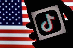 A imagem mostra a silhueta de uma mão segurando um smartphone com o logotipo do TikTok em destaque. Ao fundo, há uma bandeira dos Estados Unidos visível, criando um contraste entre a mão e o dispositivo em primeiro plano.