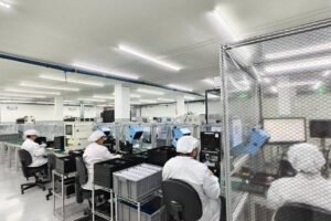 A imagem mostra um ambiente de trabalho em uma fábrica, com várias estações de trabalho. Há pessoas vestindo uniformes brancos e toucas, sentadas em cadeiras em frente a mesas com equipamentos eletrônicos. Algumas áreas estão cercadas por uma grade de proteção. O espaço é bem iluminado e organizado, com prateleiras e equipamentos visíveis ao fundo.