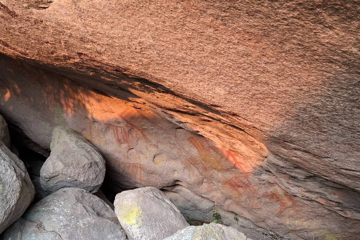 A imagem mostra uma superfície rochosa com uma coloração avermelhada, localizada sob uma saliência. Há várias pedras de diferentes tamanhos e formas ao redor da base da rocha.