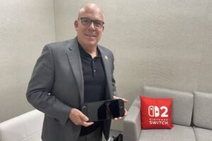 Um homem de óculos e terno cinza está em pé segurando um console Nintendo Switch 2. Ele está em um ambiente interno com paredes cinzas e um sofá claro ao fundo. Ao lado dele, há uma almofada vermelha com o logotipo do Nintendo Switch 2.