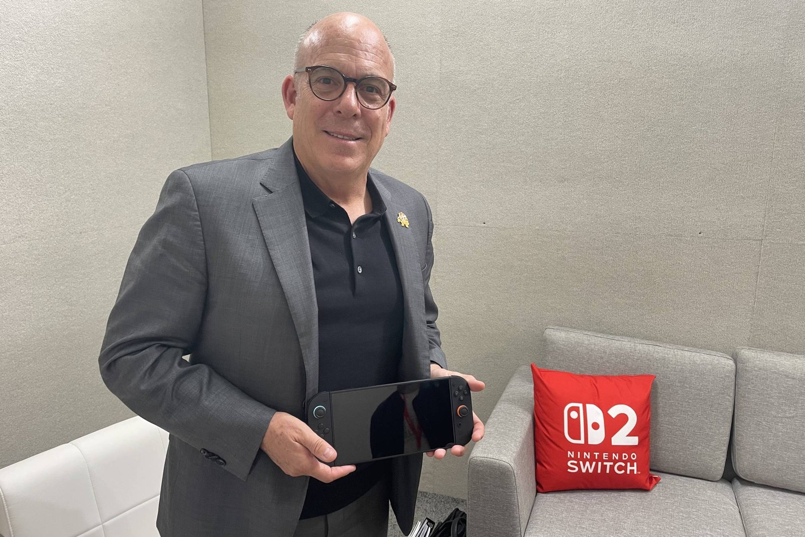 Um homem de óculos e terno cinza está em pé segurando um console Nintendo Switch 2. Ele está em um ambiente interno com paredes cinzas e um sofá claro ao fundo. Ao lado dele, há uma almofada vermelha com o logotipo do Nintendo Switch 2.