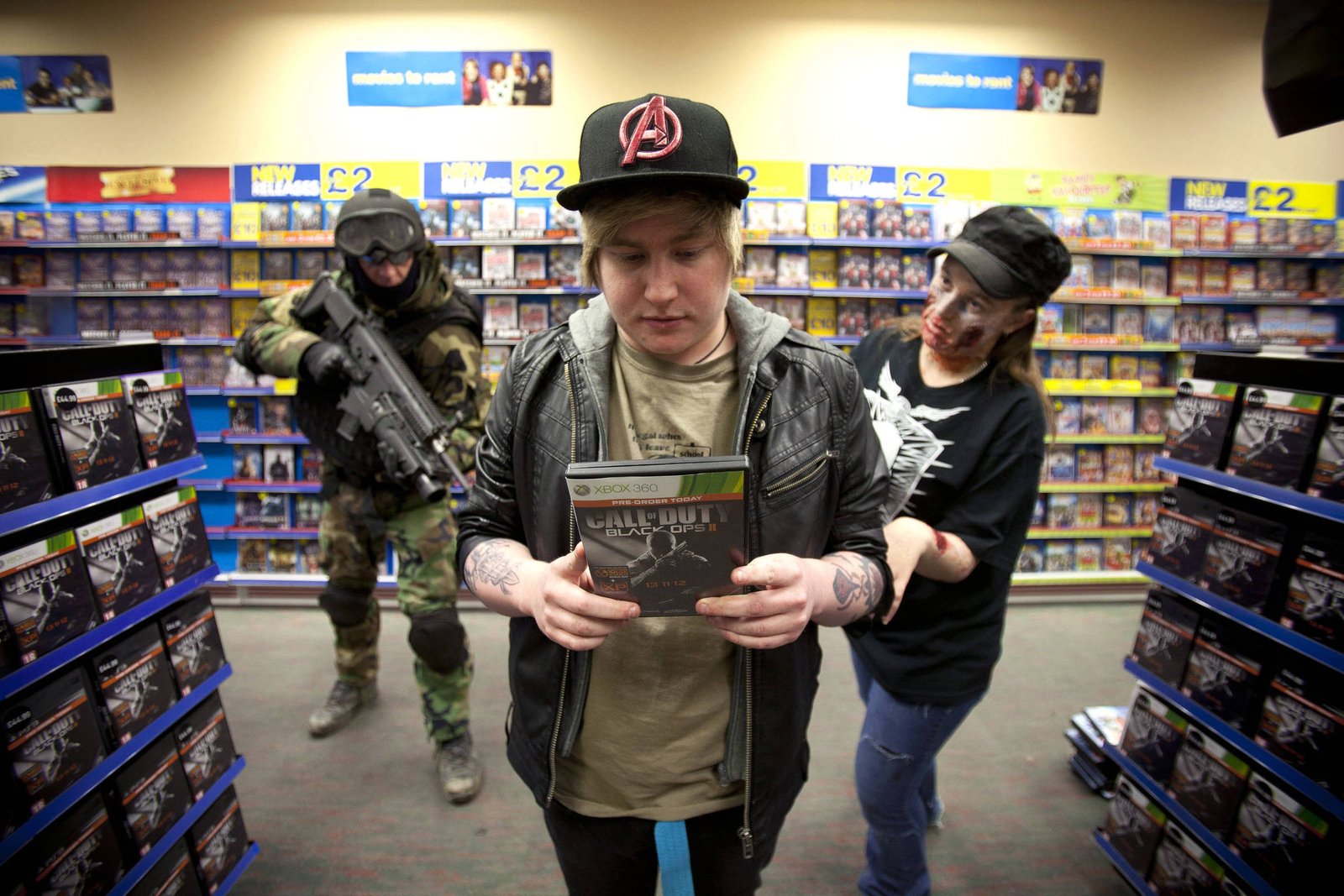 A imagem mostra uma cena em uma loja de jogos. Um jovem com cabelo loiro e um boné preto está segurando um jogo chamado