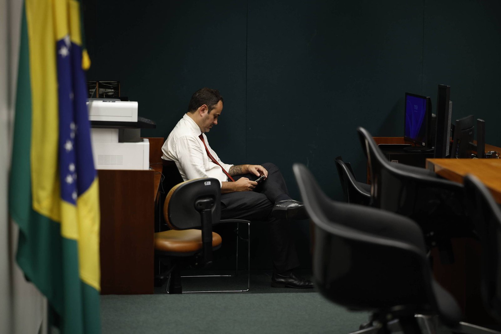 Um homem sentado em uma cadeira em um ambiente de trabalho, aparentemente concentrado em seu celular. Ao fundo, há uma bandeira do Brasil e mesas de trabalho com computadores. O ambiente é bem iluminado e possui paredes verdes.