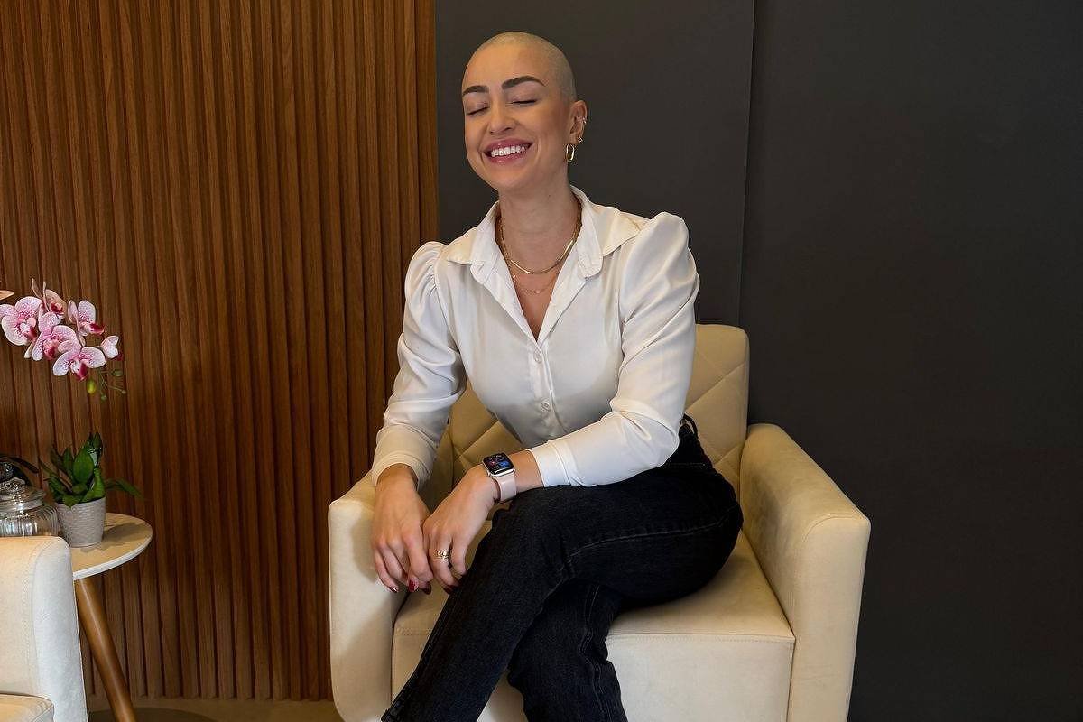 Uma mulher com cabelo raspado está sentada em uma cadeira clara, sorrindo. Ela usa uma camisa branca e calças pretas. Ao lado dela, há uma mesa pequena com um vaso de flores rosas. O fundo é composto por uma parede escura e painéis de madeira.