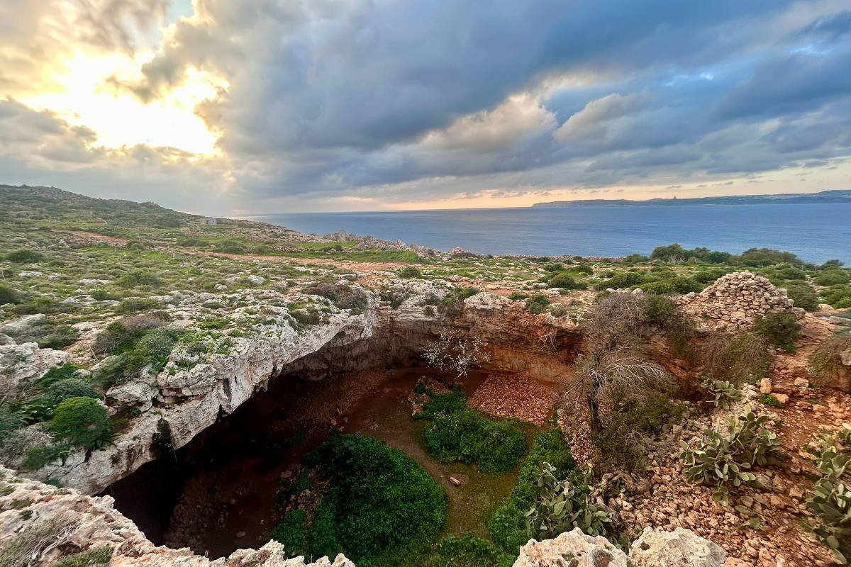 A imagem mostra uma vista panorâmica de uma falésia com vegetação ao redor, localizada em uma costa. No fundo, há um mar calmo sob um céu nublado, com nuvens iluminadas pela luz do sol que se esconde. A falésia apresenta uma abertura que parece ser uma caverna ou depressão, com vegetação verde no interior.