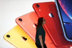 A imagem mostra uma mulher em pé ao lado de um grande painel que exibe três modelos de iPhones XR em cores vibrantes: amarelo, laranja e vermelho. O fundo é claro, destacando os dispositivos. A mulher está usando uma camiseta escura e parece estar observando os telefones.