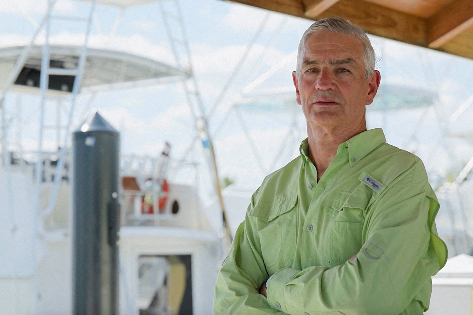 Um homem de cabelo grisalho está posicionado em primeiro plano, com os braços cruzados. Ele usa uma camisa de manga longa verde-clara. Ao fundo, é possível ver um barco de pesca e uma estrutura náutica, com um céu claro e algumas nuvens.