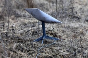 A imagem mostra uma antena de formato cônico, apoiada em uma base de metal, situada em um terreno seco e coberto por grama amarelada. O ambiente parece ser um campo aberto, com vegetação escassa ao redor. Um cabo está conectado à antena, estendendo-se pelo solo.