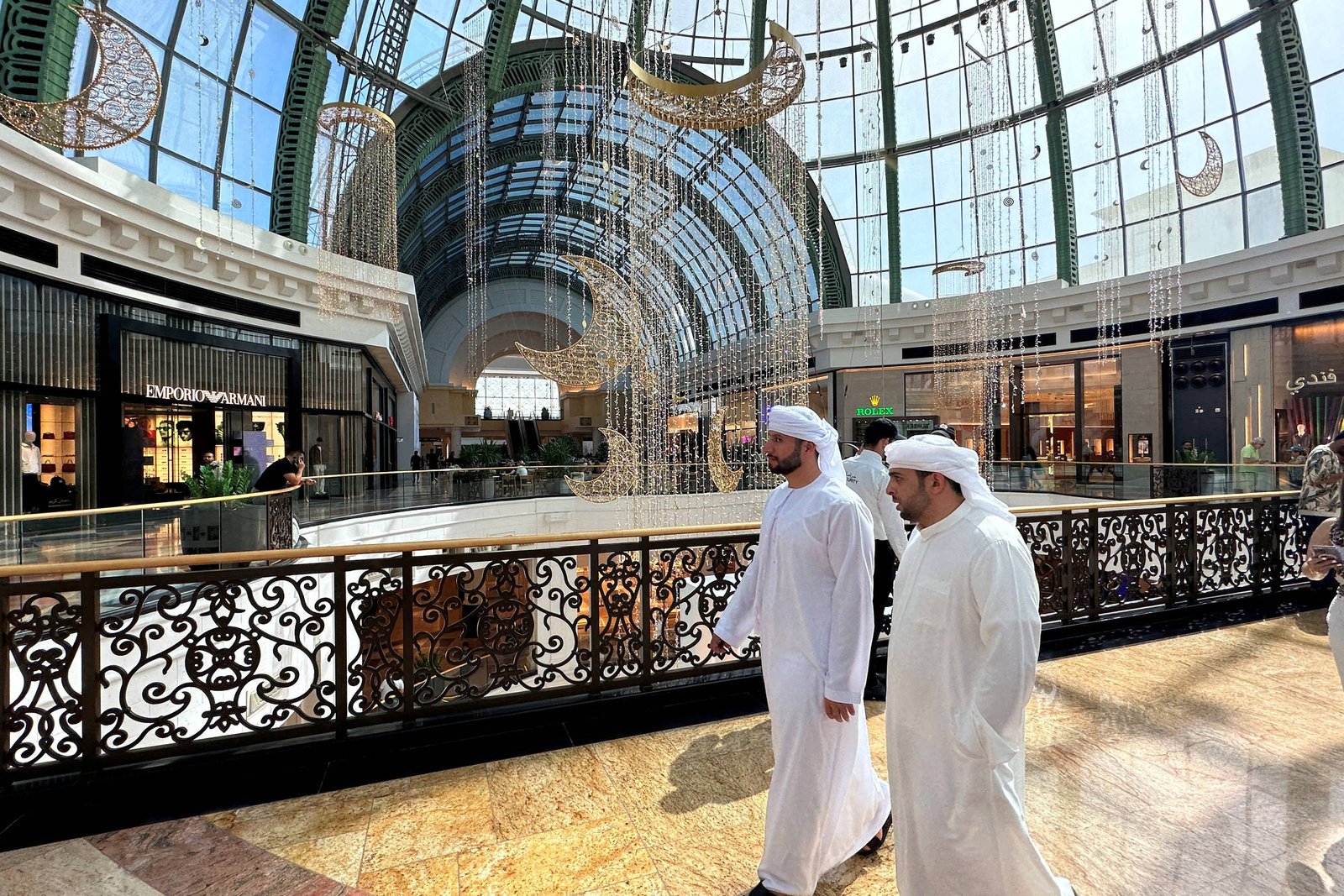 A imagem mostra o interior de um shopping em Dubai, com um grande teto de vidro que permite a entrada de luz natural. Duas pessoas vestindo roupas tradicionais brancas estão caminhando em direção à câmera, enquanto ao fundo há lojas e uma decoração elegante. O ambiente é espaçoso e moderno, com detalhes arquitetônicos sofisticados.