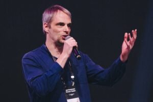 Um homem está em um palco, segurando um microfone e gesticulando com a mão direita. Ele usa uma camisa azul escura e calças claras. Ao fundo, há uma iluminação suave e um público visível, mas desfocado.