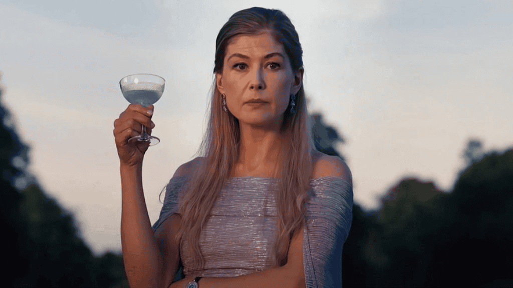 Rosamund Pike em cena de Saltburn (2023), vestindo um elegante vestido prateado ombro a ombro, segurando uma taça e olhando com expressão séria. O fundo desfocado mostra uma área externa ao entardecer