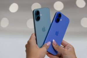 A imagem mostra duas mãos segurando dois smartphones da Apple, um na cor azul e outro na cor verde. Os dispositivos estão posicionados um ao lado do outro, com o fundo desfocado e iluminado por luzes circulares. As unhas da pessoa que segura os celulares estão pintadas de forma decorativa.