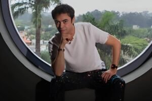 Um homem está posicionado em uma janela redonda, com uma vista de árvores e casas ao fundo. Ele usa uma camiseta branca e calças brilhantes. O ambiente é bem iluminado, com palmeiras visíveis do lado de fora.