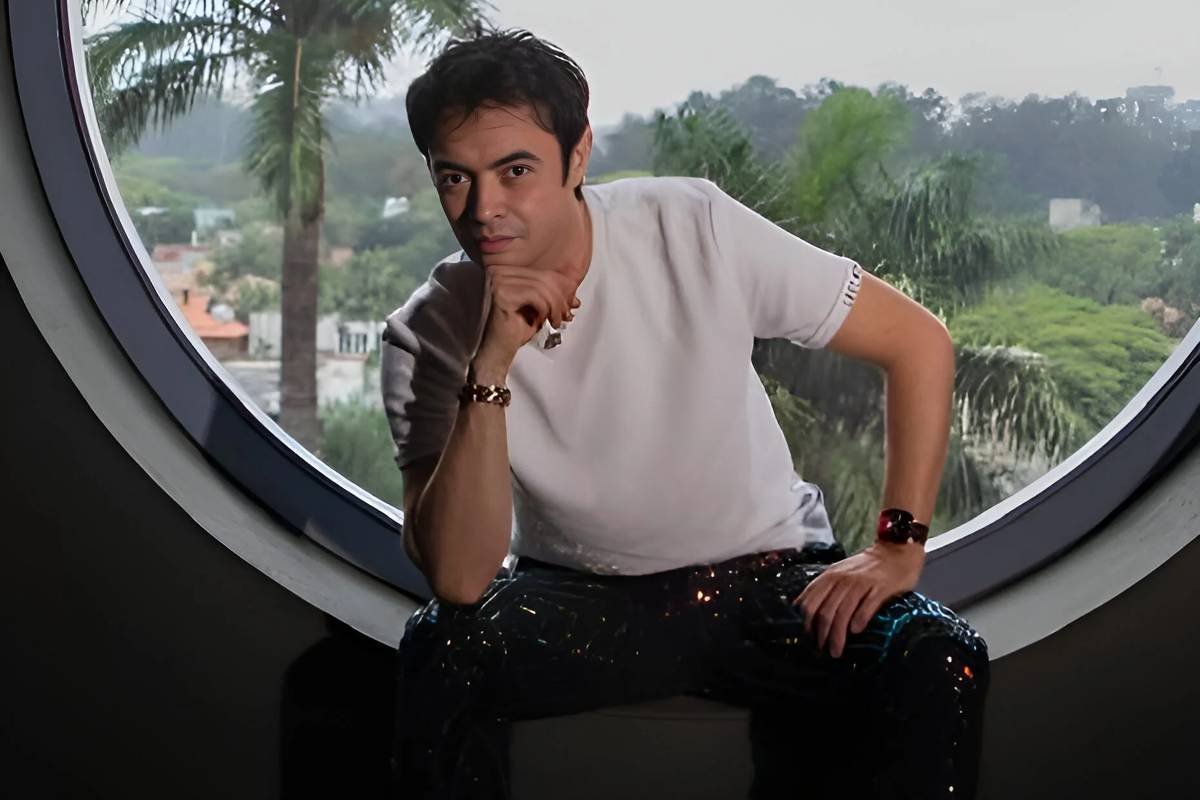 Um homem está posicionado em uma janela redonda, com uma vista de árvores e casas ao fundo. Ele usa uma camiseta branca e calças brilhantes. O ambiente é bem iluminado, com palmeiras visíveis do lado de fora.