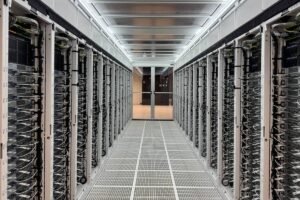 A imagem mostra um corredor de uma sala de servidores, com fileiras de servidores organizados em racks em ambos os lados. O ambiente é bem iluminado, com luzes no teto e um piso de grade. No fundo, há uma porta de vidro que leva a outra área.