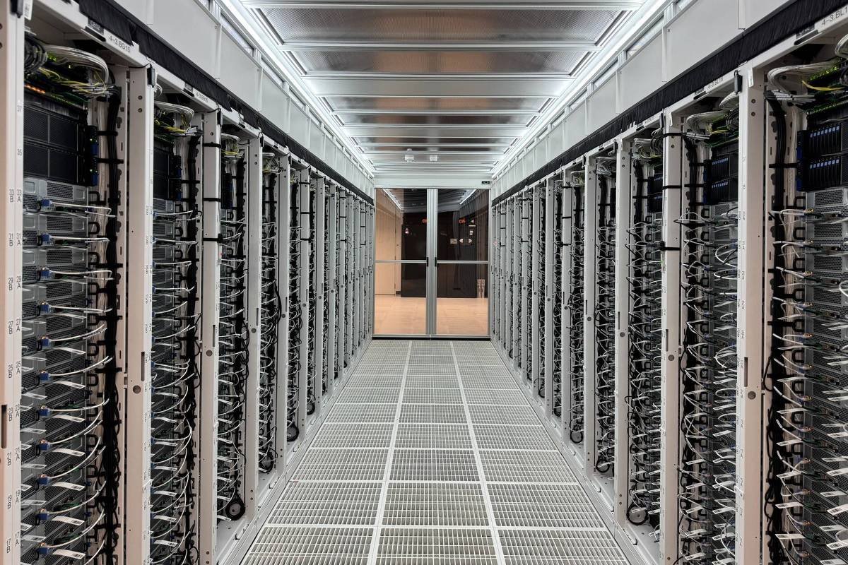 A imagem mostra um corredor de uma sala de servidores, com fileiras de servidores organizados em racks em ambos os lados. O ambiente é bem iluminado, com luzes no teto e um piso de grade. No fundo, há uma porta de vidro que leva a outra área.