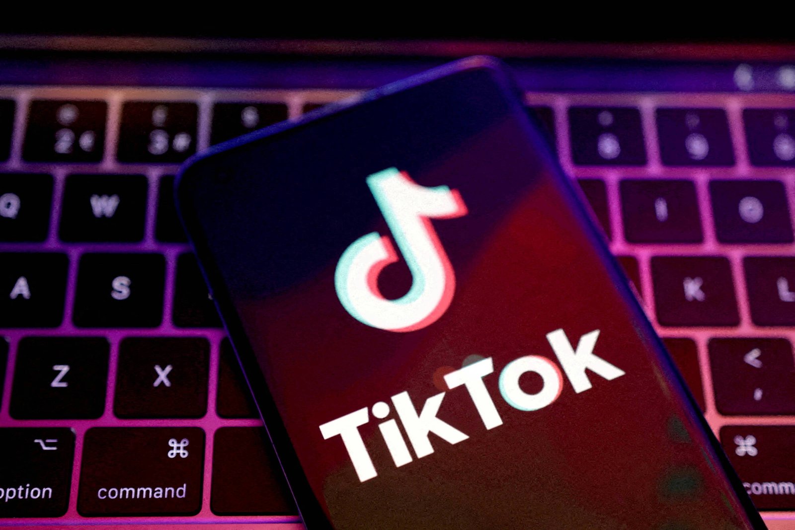 A imagem mostra um smartphone com a tela exibindo o logotipo do TikTok. O fundo é um teclado de computador, e a iluminação é suave, com tons de roxo e azul. O logotipo é composto por um símbolo estilizado e o nome