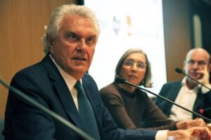 A imagem mostra um painel de conferência com três pessoas sentadas. À esquerda, um homem de cabelo grisalho e terno escuro fala ao microfone, expressando-se de forma intensa. No centro, uma mulher com cabelo liso e óculos observa atentamente. À direita, um homem de cabelo curto e camisa clara escuta. O fundo é uma parede de madeira com uma tela de apresentação visível.