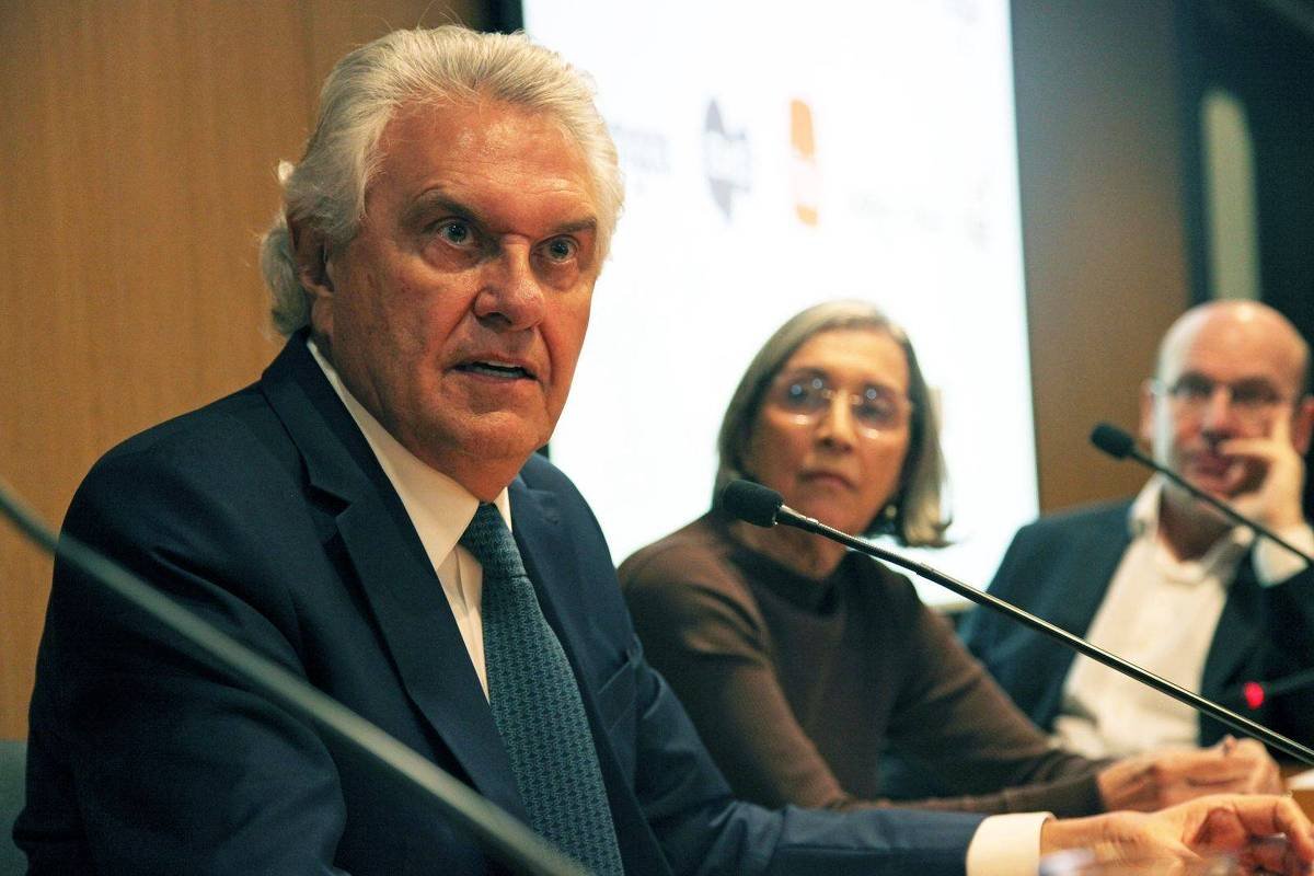 A imagem mostra um painel de conferência com três pessoas sentadas. À esquerda, um homem de cabelo grisalho e terno escuro fala ao microfone, expressando-se de forma intensa. No centro, uma mulher com cabelo liso e óculos observa atentamente. À direita, um homem de cabelo curto e camisa clara escuta. O fundo é uma parede de madeira com uma tela de apresentação visível.