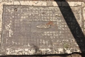 Uma placa de concreto retangular, com uma superfície desgastada e suja. A placa possui um desenho ou símbolo no centro, que parece ser um emblema ou logotipo, e está cercada por uma moldura. A iluminação da imagem sugere que há uma sombra projetada sobre a placa, possivelmente de um objeto ou pessoa acima dela.