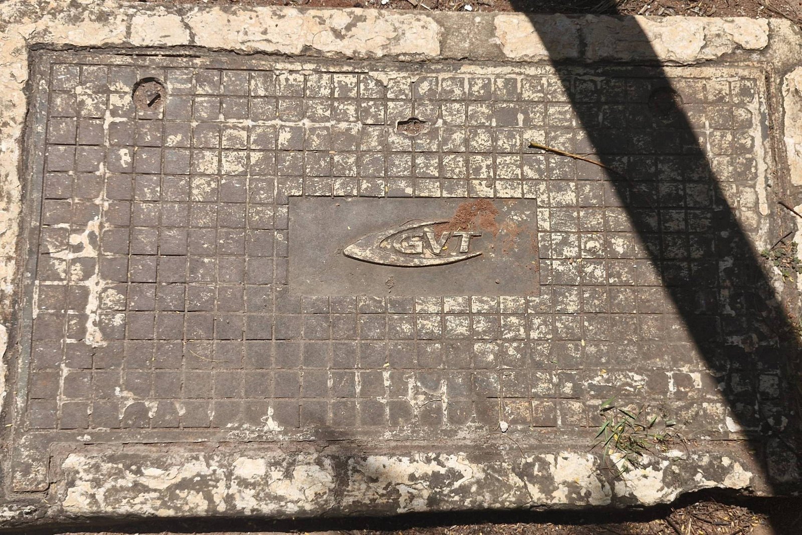 Uma placa de concreto retangular, com uma superfície desgastada e suja. A placa possui um desenho ou símbolo no centro, que parece ser um emblema ou logotipo, e está cercada por uma moldura. A iluminação da imagem sugere que há uma sombra projetada sobre a placa, possivelmente de um objeto ou pessoa acima dela.