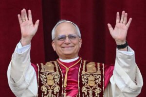 Um homem com cabelos grisalhos e óculos, vestindo uma túnica branca com detalhes em vermelho e dourado, está sorrindo e levantando as mãos em um gesto de saudação. O fundo é de um tecido vermelho