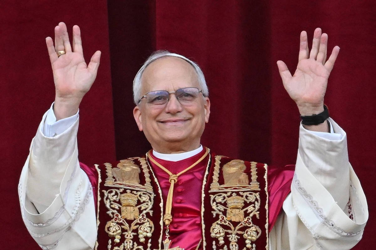 Um homem com cabelos grisalhos e óculos, vestindo uma túnica branca com detalhes em vermelho e dourado, está sorrindo e levantando as mãos em um gesto de saudação. O fundo é de um tecido vermelho