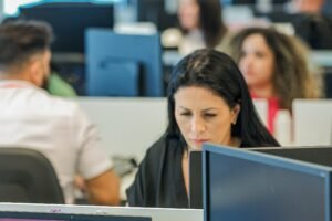 A imagem mostra um ambiente de escritório moderno, com várias pessoas trabalhando em computadores. Em primeiro plano, uma mulher com cabelo longo e escuro está concentrada em seu trabalho, olhando para a tela do computador. Ao fundo, outras pessoas também estão visíveis, algumas de costas, em um espaço de trabalho compartilhado.