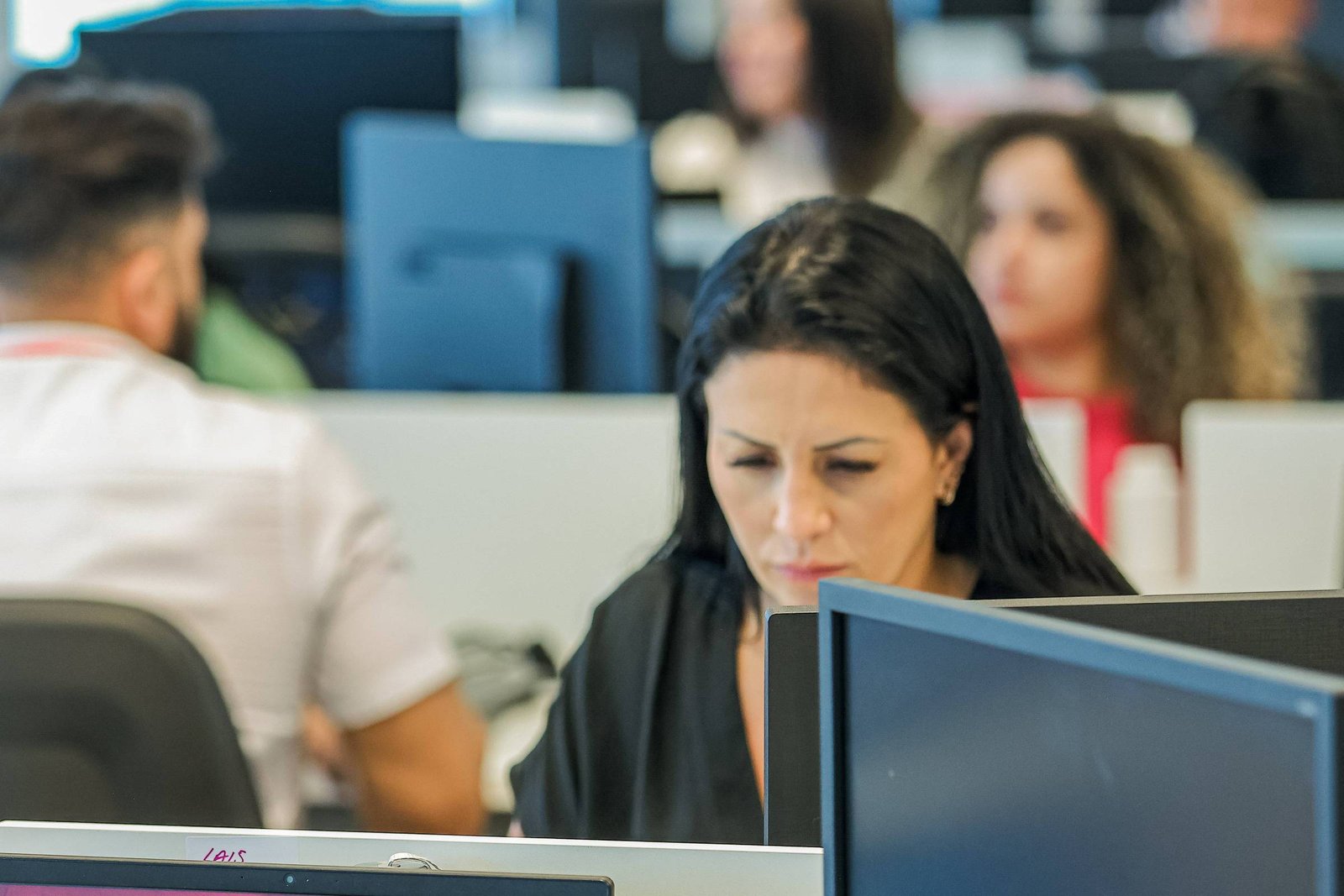 A imagem mostra um ambiente de escritório moderno, com várias pessoas trabalhando em computadores. Em primeiro plano, uma mulher com cabelo longo e escuro está concentrada em seu trabalho, olhando para a tela do computador. Ao fundo, outras pessoas também estão visíveis, algumas de costas, em um espaço de trabalho compartilhado.