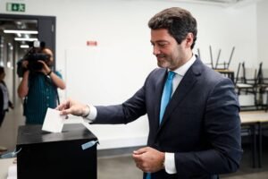Um homem de terno escuro e gravata azul está colocando um papel em uma urna de votação. Ele parece concentrado e está sorrindo levemente. Ao fundo, um fotógrafo está registrando o momento. O ambiente é claro e moderno, com mesas e cadeiras visíveis.
