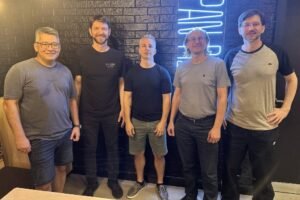 Cinco homens estão posando para a foto em um ambiente interno. Eles estão em pé, alinhados, com sorrisos. O homem à esquerda usa óculos e uma camiseta cinza, o segundo usa uma camiseta preta, o terceiro está com uma camiseta preta e shorts claros, o quarto usa uma camiseta clara e o quinto está com uma camiseta cinza escura