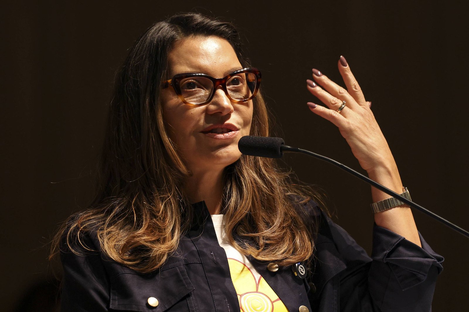 Uma mulher com cabelos longos e ondulados, usando óculos e uma blusa branca com detalhes coloridos, está falando em um microfone. Ela gesticula com a mão direita, enquanto a mão esquerda está apoiada no microfone. O fundo é escuro, sugerindo um ambiente de palestra ou conferência.