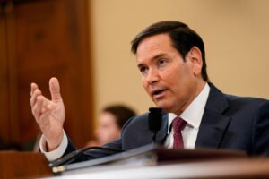 Um homem de terno escuro está falando em um microfone durante uma audiência no Congresso. Ele gesticula com a mão esquerda enquanto se dirige a um público fora do foco. Ao fundo, é possível ver outras pessoas, mas não estão claramente visíveis.