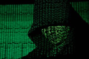 A imagem mostra a silhueta de uma figura encapuzada, com um fundo repleto de códigos binários em verde. A figura parece estar em um ambiente digital, transmitindo uma sensação de mistério e tecnologia.