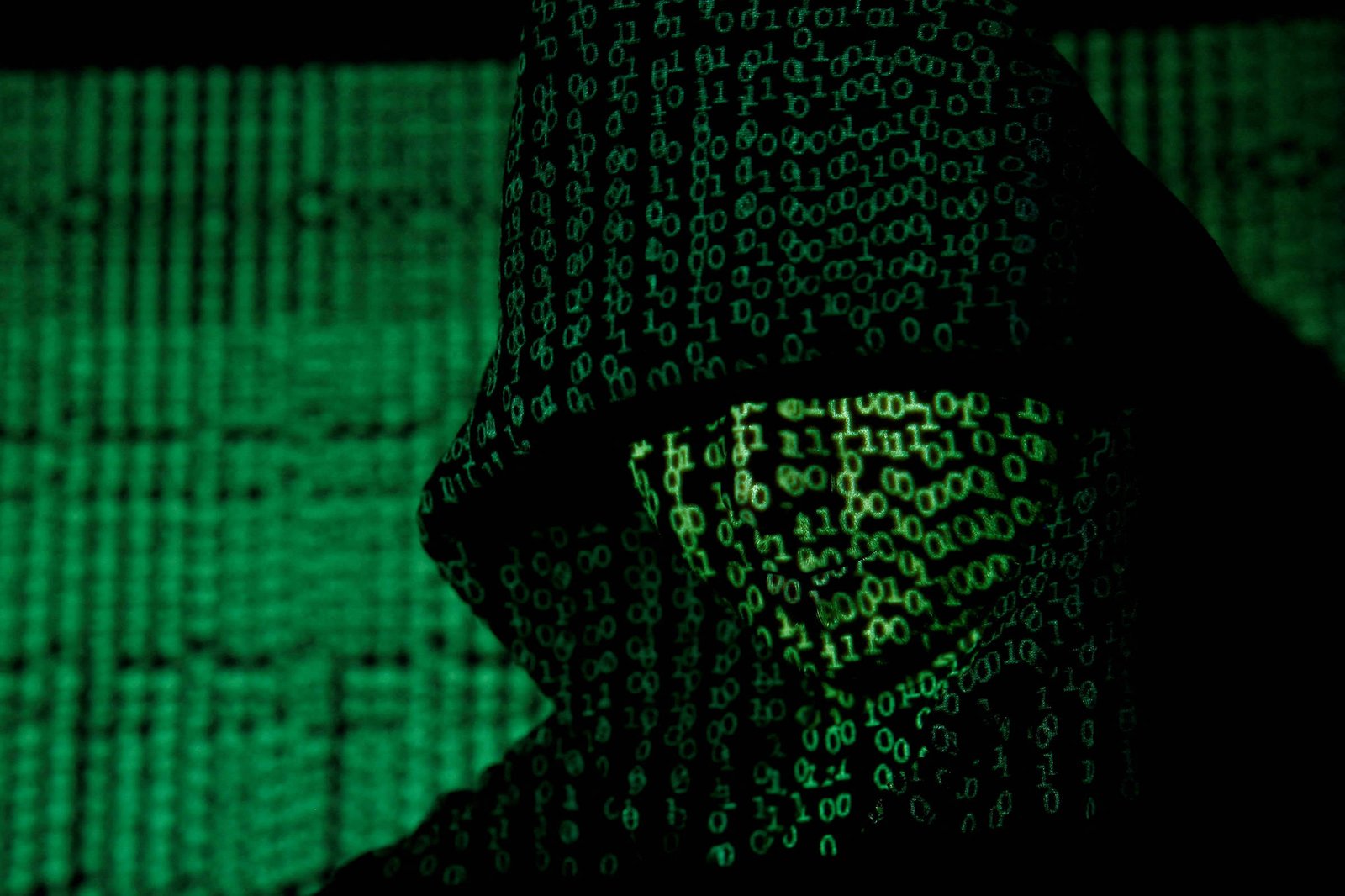 A imagem mostra a silhueta de uma figura encapuzada, com um fundo repleto de códigos binários em verde. A figura parece estar em um ambiente digital, transmitindo uma sensação de mistério e tecnologia.