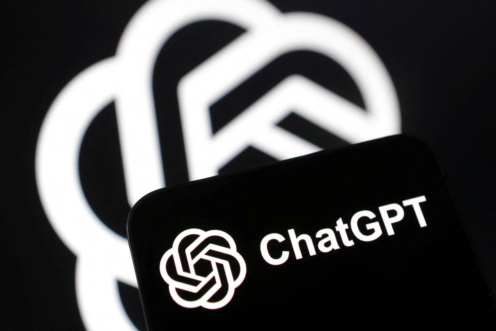 A imagem mostra um smartphone com o logotipo do ChatGPT em destaque na tela. O fundo é escuro, com um padrão de logotipo em um tom mais claro, criando um contraste. O logotipo do ChatGPT é composto por um símbolo estilizado e o texto