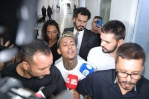 A imagem mostra um grupo de pessoas em um corredor, onde um homem com cabelo curto e tatuagens está sendo cercado por repórteres e câmeras. Ele parece estar falando, enquanto outros indivíduos ao seu redor observam. Há uma mulher e vários homens, alguns com microfones e câmeras, capturando a cena. O ambiente é bem iluminado e moderno, com paredes brancas e um piso claro.